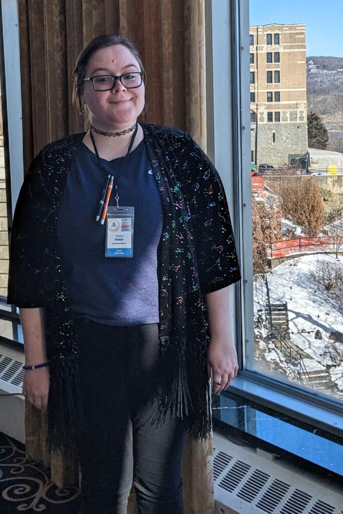 Stoker smiling and standing in front of a window.  Around their neck is a lanyard with a two pens and a badge for a conference.