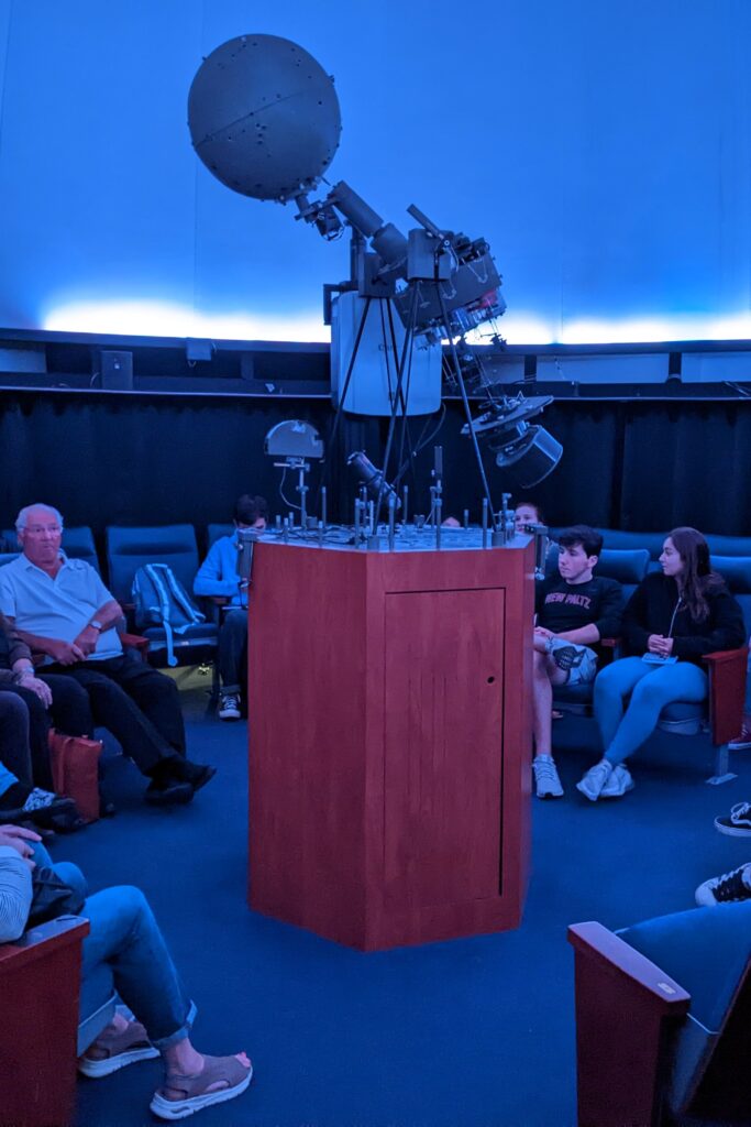 The lit interior of the John R. Kirk Planetarium with the spherical Spitz A3P "Starball" projector at the center and people sitting in the seats around it.